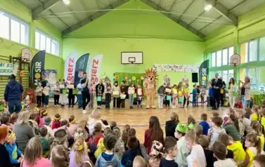Duża grupa dzieci na sali gimnastycznej w szkole. Jedni siedzą na krzesełkach inni występują przed nimi z maskątką miasta Zgierza- osoba przebrana za jeża