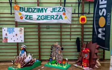 3 kukiełki jeża stojące w sali gimnastycznej pod napisem Budzimy jeża ze Zgierza