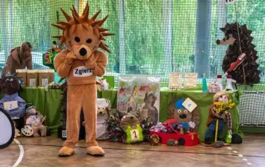 Jeż - maskotka Zgierza (osoba w beżowym kostiumie jeża) stoi na tle stolik&oacute;w z ustawionymi jeżami kukiełkami przygotowany na konkur
