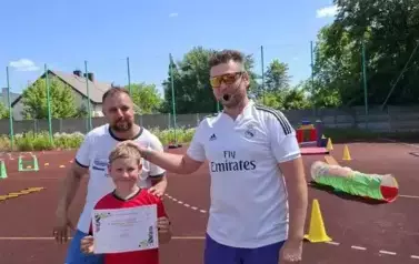 Na tle boiska szkolnego z torem przeszk&oacute;d stoi dw&oacute;ch opiekun&oacute;w w białych koszulkach i niebieskich spodenkach. przed nimi stoi chłopiec w czerwonej koszulce i zielonych spodenkach, trzymający przed sobą dyplom za udział w szkolnym dniu sportu i profilaktyk