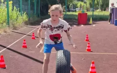 Na boisku szkolnym dziewczynka w białej bluzce z nadrukiem i niebieskich spodenkach toczy przed sobą czarną oponę.
