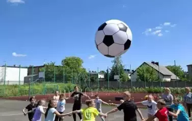 Grupa uczni&oacute;w na boisku szkolnym, ustawiona w kole trzyma się za ręce i podrrzuca wielką biało czarną piłkę