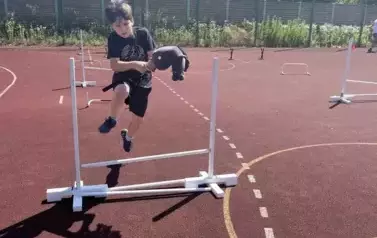 Chłopiec w czarny stoju sportowym, z głową konia na kiju między nogami pokonuje tor przeszk&oacute;d na boisku szkolnym - skacze przez płotki