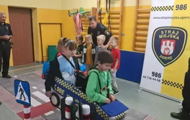 Grupa uczni&oacute;w pokonuje tor ruchu drogowego. Dw&oacute;jka dzieci ma makietę aut. Pan i pani w mundurach Straży Miejskiej nadzorują prawidłowe wykonanie zadań.