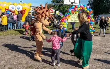 na tle kolorowej bramy z balon&oacute;w i rozłożonej zjeżdżalni na boisku szkolnym tańczy w k&oacute;łku trzymając się za ręce: troje dzieci, nauczycielka i postać przebrana za jeża. 