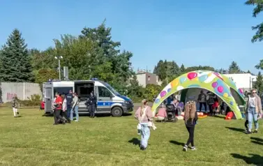 Na boisku szkolnym ustawiony namiot pod kt&oacute;ry ułozono materace do pokaz&oacute;w udzielania pierwszej pomocy. Po lewej stronie namiotu stoi radiow&oacute;z policyjny z otwrtymi bocznymi drzwiami. Przy namiocie i radiowozie grupy dzieci i dorosłych.