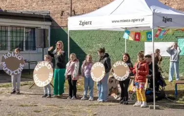 Pod namiotem ustawionym na boisku przed szkołą stoi grupa dzieci trzymająca przed sobą koła z szarej tektury z napisem koło muzyczne. Jedna dziewczynka trzyma mikrofon i śpiewa. Nauczycielka pomiędzy nimi z podniesionymi rękami zachęca do zabawy. Za nimi 