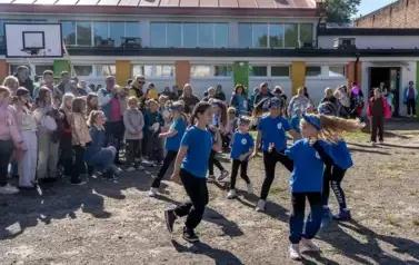 Na boisku szkolnym sześć dziewczynek w niebieskich koszulkach i czarnych getrach tańczy w kole. Wok&oacute;ł nich duża grupa obserwujących występ.
