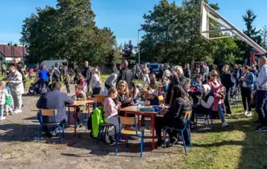 Duża grupa dzieci i dorosłych siedzi przy stolikach ustawionych na boisku szkolnym. W tle spacerujący dorośli i dzieci.