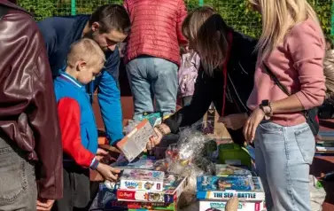 Na stolikach ustawionych na boisku szkolnym ułożone do sprzedaży gry planszowe. Po lewej stronie stolika chłopiec w kolorwej bluzie z rodzicem w szarej kurtce oglądają gry. Po prawej stronie stoją dwie nauczycielki sprzedające: na pierwszym planie blondyn