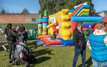 Na boisku szkolnym ustawiona kolorowa, dmuchana zjeżdzalnia dla dzieci. Wok&oacute;ł grupa dorosłych i dzieci, przygląda się zabawom.