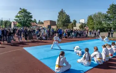 Na boisku szkolnym ułożona niebieska mata, na kt&oacute;rej grupa dzieci ćwiczących karate wykonuje pokazy ćwiczeń. Wszystkie w białych strojach karatek&oacute;w. W tle duża grupa dorosłych i dzieci przygląda się pokazom. 