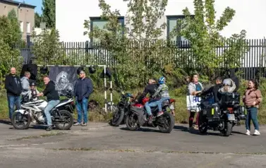 Na boisku szkolnym grupa trzech motocyklist&oacute;w prezentuje swoje motory. Do przejażdzki na jednym z nich przygotowuje się dziewczynka w kolorowym kasku na głowie. Duża grupa dzieci i dorosłych przygląda się.