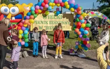 W wielkim łuku z kolorowych balon&oacute;w stojącym na szkolnym boisku wisi napis Wielki piknik rodzinny i stoi tr&oacute;jka dzieci pozująca do zdjęć: od lewej chłopiec w szarej kurtce i niebieskich spodniach, w środku dziewczynka w r&oacute;żowej kurtce i szarych leginsach,