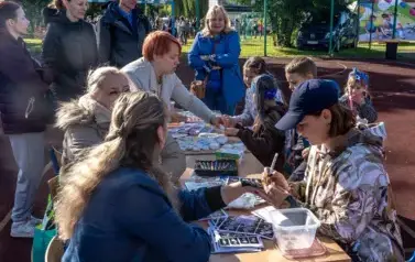 Przy trzech stolikach ustawionych na boisku szkolnym podczas pikniku rodzinnego, siedzą trzy panie wykonujące tatuaże. Grupa dzieci samodzielnie koloruje figurki. Wok&oacute;ł stolika przygląda się dzieciom trzy panie i jeden pan. 