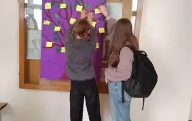 Dwie dziewczynki przyklejają na plakacie z narysowanym drzewem i powieszonym na ścianie korytarza, karteczki z dobrymi słowami. Stoją tyłem. Jedna w szarej bluzie i czarnych spodniach, druga w szarej bluzie, niebieskich spodniach z czrnym tornistrem na pl
