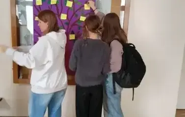 Trzy dziewczynki przyklejają na plakacie z narysowanym drzewem i powieszonym na ścianie korytarza, karteczki z dobrymi słowami. Stoją tyłem. Od lewej: jedna w szarej bluzie i niebieskich spodniach, w środku w szarej bluzie i czarnych spodniach, po prawej 