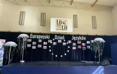 W sali gimnastycznej ustawiony podest, na kt&oacute;rym stoją kolorowe parasole: po lewej 3 r&oacute;żnej wielkości, po lewej jeden duży. Na ścianie w tle czarny materiał z napisem Dzień język&oacute;w obcych i kolorowymi flagami r&oacute;żnych państw.