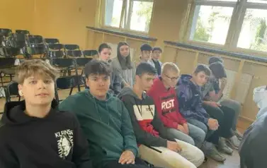 Grupa uczni&oacute;w w r&oacute;żnych strojach siedząca na czarnych krzesełkach ustwionych w rzędzie, w slai gimnastycznej. Oglądają przygotowaną imprezę z okazji Dnia język&oacute;w obcych.