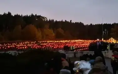 Na polanie w Puszczy Kampinowskiej, na cmentarzu leśnym paląca się o zmierzchu duża ilość zniczy. Wok&oacute;ł sylwetki os&oacute;b. 