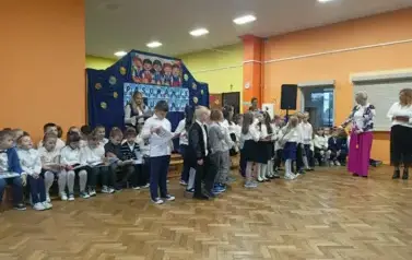 Duża grupa uczni&oacute;w klas pierwszych w strojach galowych, stoi w sali gimnastycznej podczas występ&oacute;w. W tle dekoracja: na granotowym tle napis: pasowanie na ucznia i grafika uśmiechniętych dzieci.
