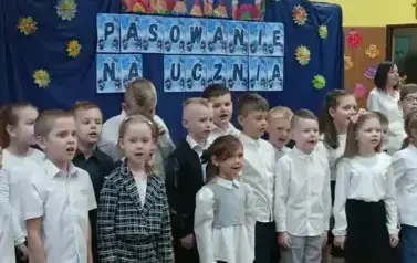 Duża grupa uczni&oacute;w klas pierwszych w strojach galowych, podczas występu w sali gimnastycznej. W tle dekoracja: na granatowym tle napis: pasowanie na ucznia i grafika uśmiechniętych dzieci.