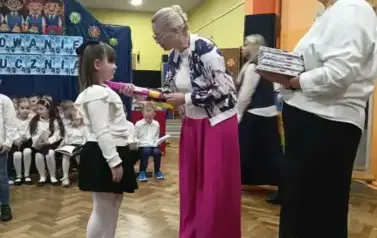 Dyrektor w fioletowych szerokich spodniach i białej bluzce pasuje na ucznia dziewczynkę, ubraną w białą bluzkę i czarną sp&oacute;dniczkę. Dotyka ramienia dziewczynki dużym fioletowym oł&oacute;wkiem.w Tle na ławeczkach siedzi grupa uczni&oacute;w klas pierwszych w strojach g