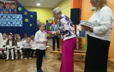 Dyrektor w fioletowych szerokich spodniach i białej bluzce pasuje na ucznia chłopca, ubranego w białą koszulę i czarne spodnie. Dotyka ramienia chłopca dużym fioletowym oł&oacute;wkiem.w Tle na ławeczkach siedzi grupa uczni&oacute;w klas pierwszych w strojach galowych,