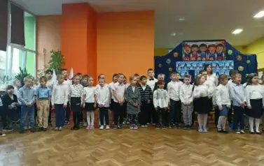 Duża grupa uczni&oacute;w klas pierwszych w strojach galowych, stoi w sali gimnastycznej podczas ślubowania. W tle dekoracja: na granotowym tle napis: pasowanie na ucznia i grafika uśmiechniętych dzieci.