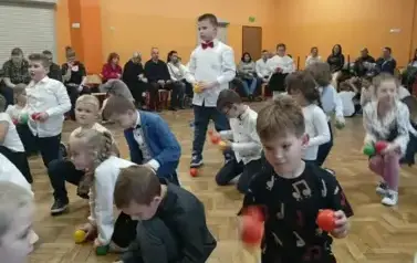 Duża grupa uczni&oacute;w klas pierwszych w strojach galowych, podczas zabawy w sali gimnastycznej. Dzieci siedzą z kolorowymi piłkami na całej powierchni sali.