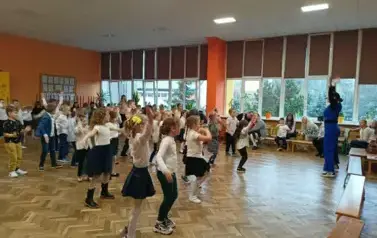 Duża grupa uczni&oacute;w klas pierwszych w strojach galowych, podczas zabawy w sali gimnastycznej. Dzieci rozstawione po całej sali tańczą, powtarzając gesty pani ubranej w niebieski str&oacute;j..