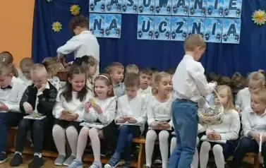 Duża grupa uczni&oacute;w klas pierwszych w strojach galowych, siedzi na ławeczkach w sali gimnastycznej. W tle dekoracja: na granotowym tle napis: pasowanie na ucznia i grafika uśmiechniętych dzieci.