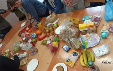 Uczniowie w sali lekcyjnej siedzą przy stolikach, na kt&oacute;rych rozłożono r&oacute;żne produkty (owoce, warzywa, soki, serki, pieczywo chrupkie, inne) do przygotowania zdrowego śniadania
