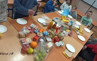 Uczniowie w sali lekcyjnej siedzą przy stolikach, na kt&oacute;rych rozłożono r&oacute;żne produkty (owoce, warzywa, soki, serki, pieczywo chrupkie, inne) do przygotowania zdrowego śniadania