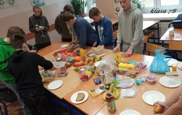 Uczniowie w sali lekcyjnej siedzą przy stolikach, na kt&oacute;rych rozłożono r&oacute;żne produkty (owoce, warzywa, soki, serki, pieczywo chrupkie, inne) do przygotowania zdrowego śniadania
