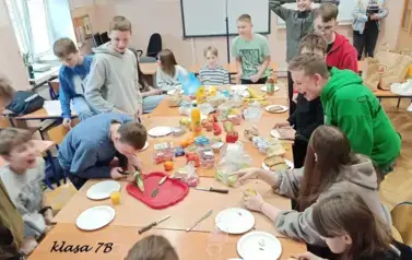 Uczniowie w sali lekcyjnej siedzą przy stolikach, na kt&oacute;rych rozłożono r&oacute;żne produkty (owoce, warzywa, soki, serki, pieczywo chrupkie, inne) do przygotowania zdrowego śniadania
