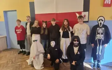 Grupa uczni&oacute;w w sali lekcyjnej w przebraniach halloweenowych pozuje do zdjęcia.