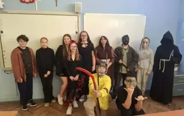 Grupa uczni&oacute;w w sali lekcyjnej w przebraniach halloweenowych pozuje do zdjęcia. 