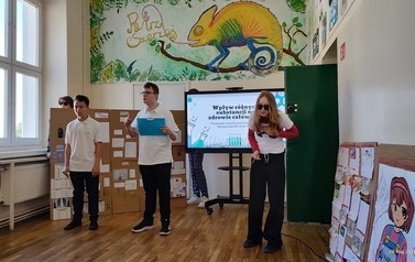 Troje uczni&oacute;w stoi na tle ektranu monitora z wyświetlonym tytułem prezentowanego projektu. Obok monitora duża makieta ze środkami uzależniającymi i ich opisem. 