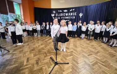 Duża grupa uczni&oacute;w klas 1 w sali gimnastycznej podczas występu i ślubowania. w tle na czarnym materiale napis Slubowanie klas pierwszych