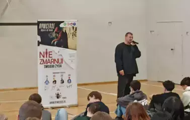 Muzyk-raper w czarnej koszulce w sali gimnastycznej, podczas występu na tle baneru z napisami RYMcerze - niezmarnuj swojego życia i zdjęć reklamujących zesp&oacute;ł. Na pierwszym planie głowy uczni&oacute;w od tyłu siedzących i oglądających spektakl.