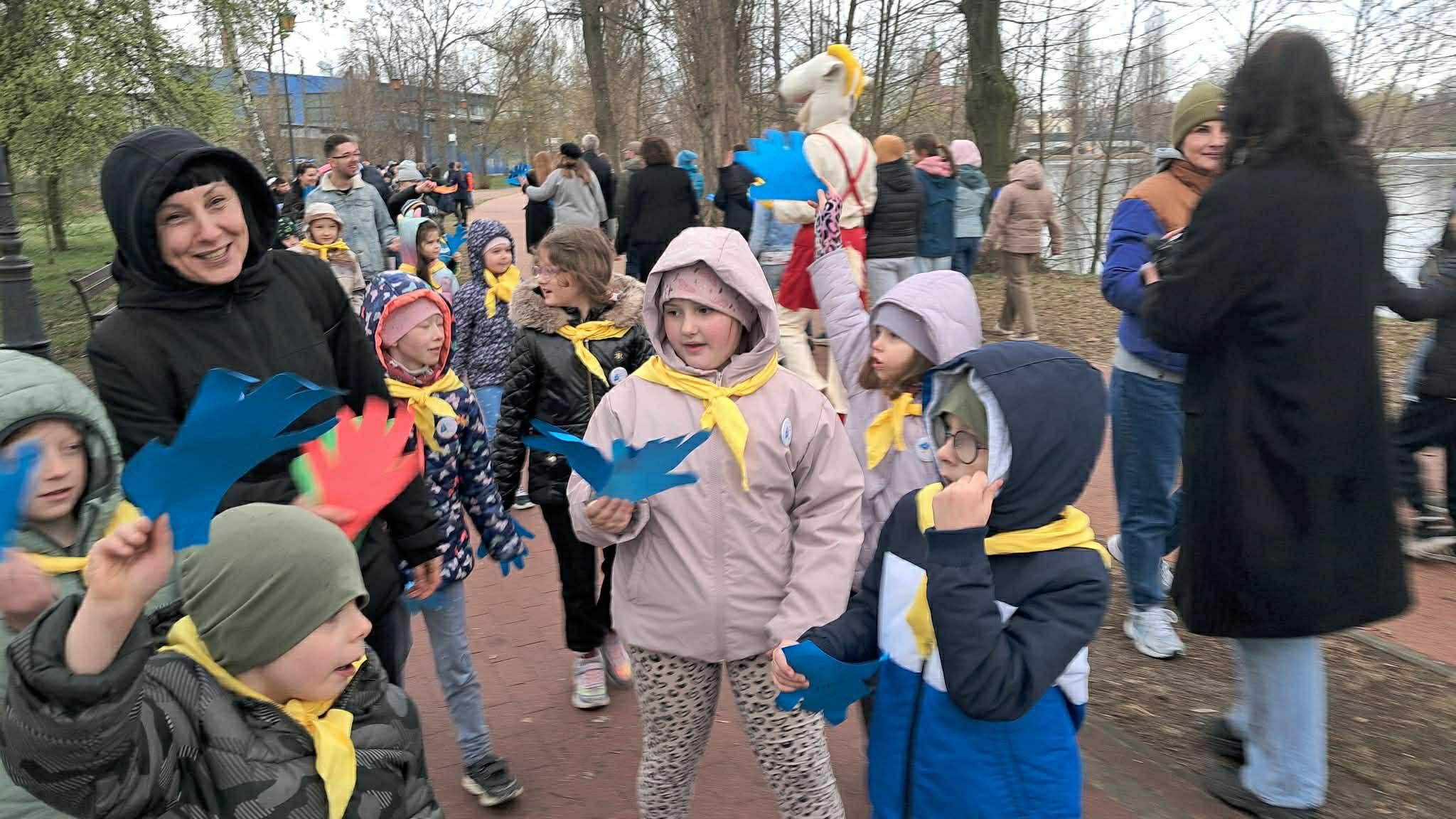 Grupa dzieci w&nbsp;parku miejskim w&nbsp;czasie obchod&oacute;w dnia autyzmu. Dzieci trzymają w&nbsp;dłoniach niebieskie łapki, a&nbsp;na szyjach mają z&oacute;łte apaszki