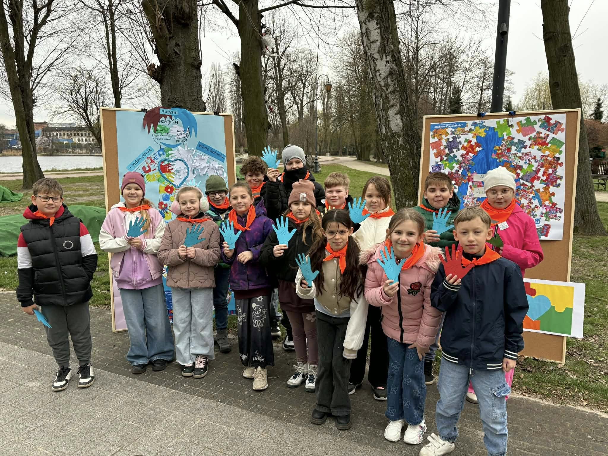 Grupa dzieci w&nbsp;parku miejskim podczas obchod&oacute;w dnia autyzmu. Dzieci mają niebieskie łapki z&nbsp;karton&oacute;w w&nbsp;dłoniach i&nbsp;ż&oacute;łte apaszki na&nbsp;szyi. Stoją przed banerami z&nbsp;informacjami o&nbsp;święcie