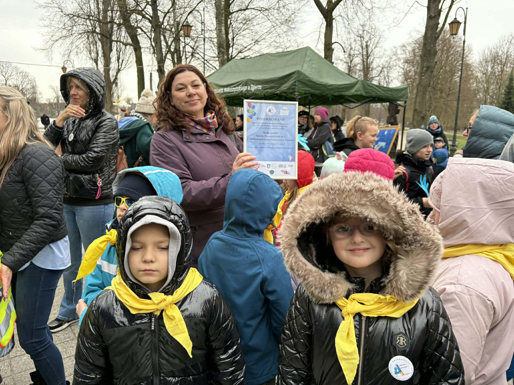 Grupa dzieci w&nbsp;parku miejskim podczas obchod&oacute;w dnia autyzmu. Dzieci maja ż&oacute;łte apaszki na&nbsp;szyjach, a&nbsp;nauczycielka dyplom z&nbsp;podziekowaniami.