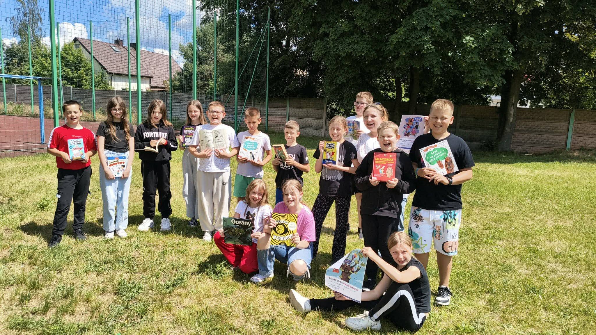 Grupa uczni&oacute;w na&nbsp;trawniku przed boiskiem szkolnym prezentuje książki do czytania