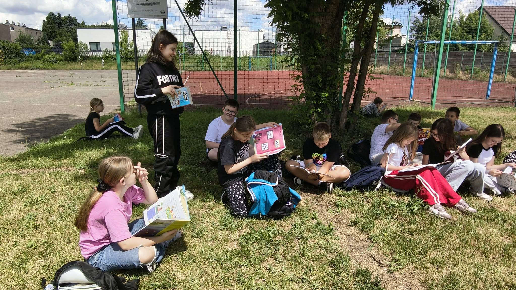 Grupa uczni&oacute;w pod drzewem na&nbsp;trawniku przed boiskiem szkolnym czyta książki