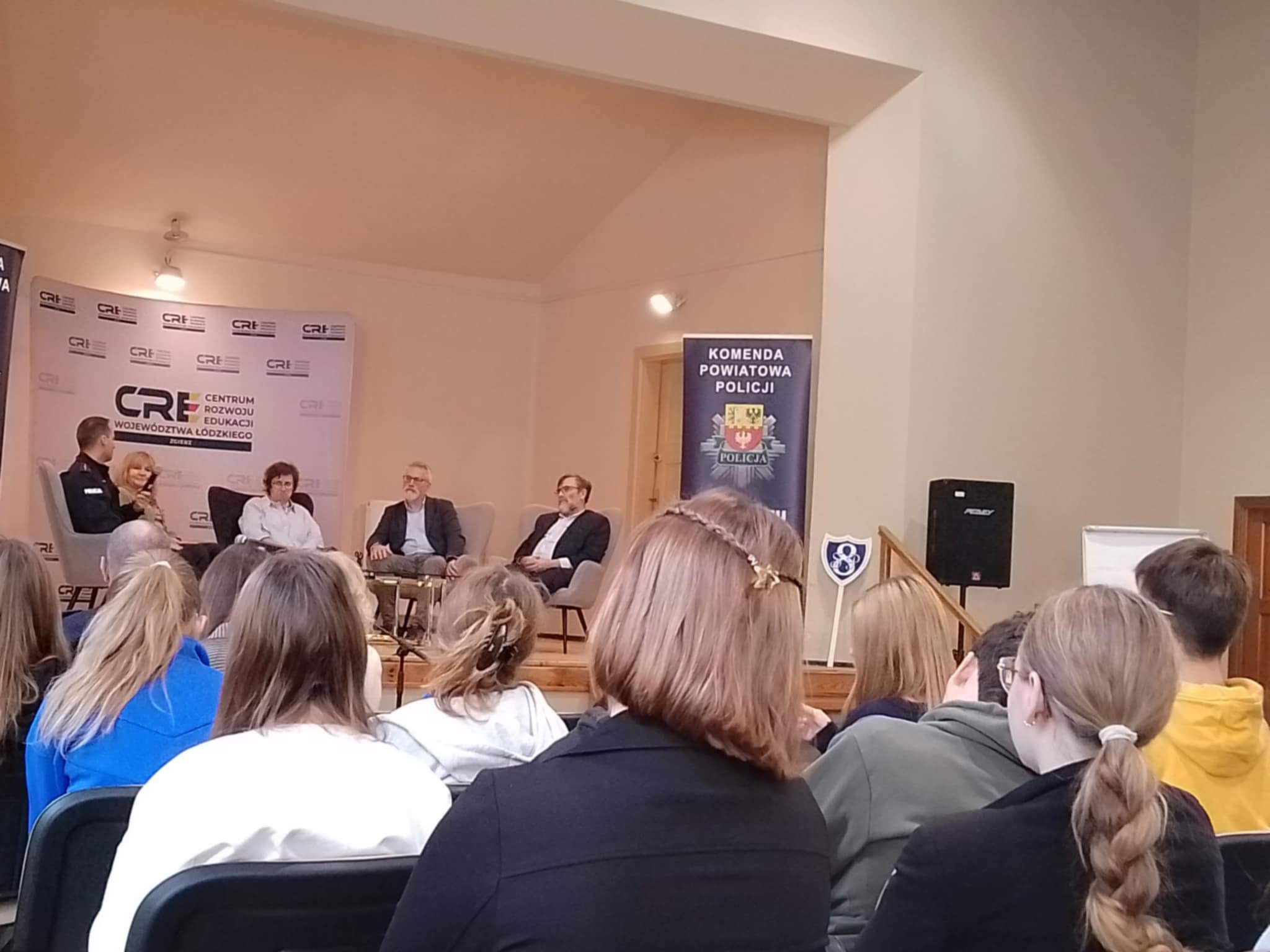 Grupa młodzieży i&nbsp;dorosłych siedzi na&nbsp;krzesłach, słuchając osoby siedzące na&nbsp;scenie. Na scenie siedzą: od lewej: policjant, dwie panie w&nbsp;białych bluzkach i&nbsp;ciemnych sp&oacute;dnicach, 2 pan&oacute;w w&nbsp;garniturach. Po prawej baner informujący o&nbsp;wydarzeniu - debacie.