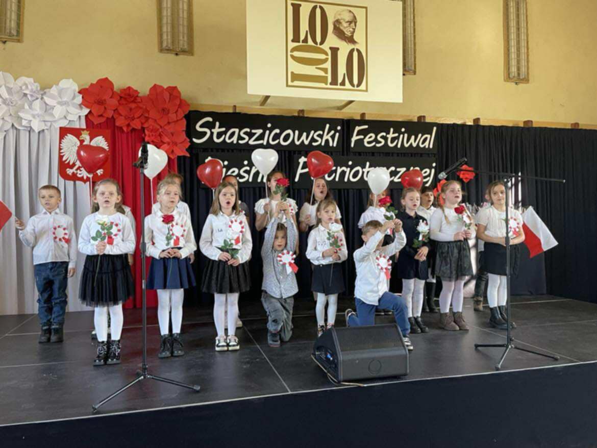 Grupa uczni&oacute;w w&nbsp;galowych strojach z&nbsp;czerwonymi r&oacute;żami w&nbsp;dłoniach podczas występu na&nbsp;sali gimnastycznej na&nbsp;Festiwalu puieśni patriotycznej. W tle informacje o&nbsp;festiwalu i&nbsp;logo liceum im. S. Staszicae 