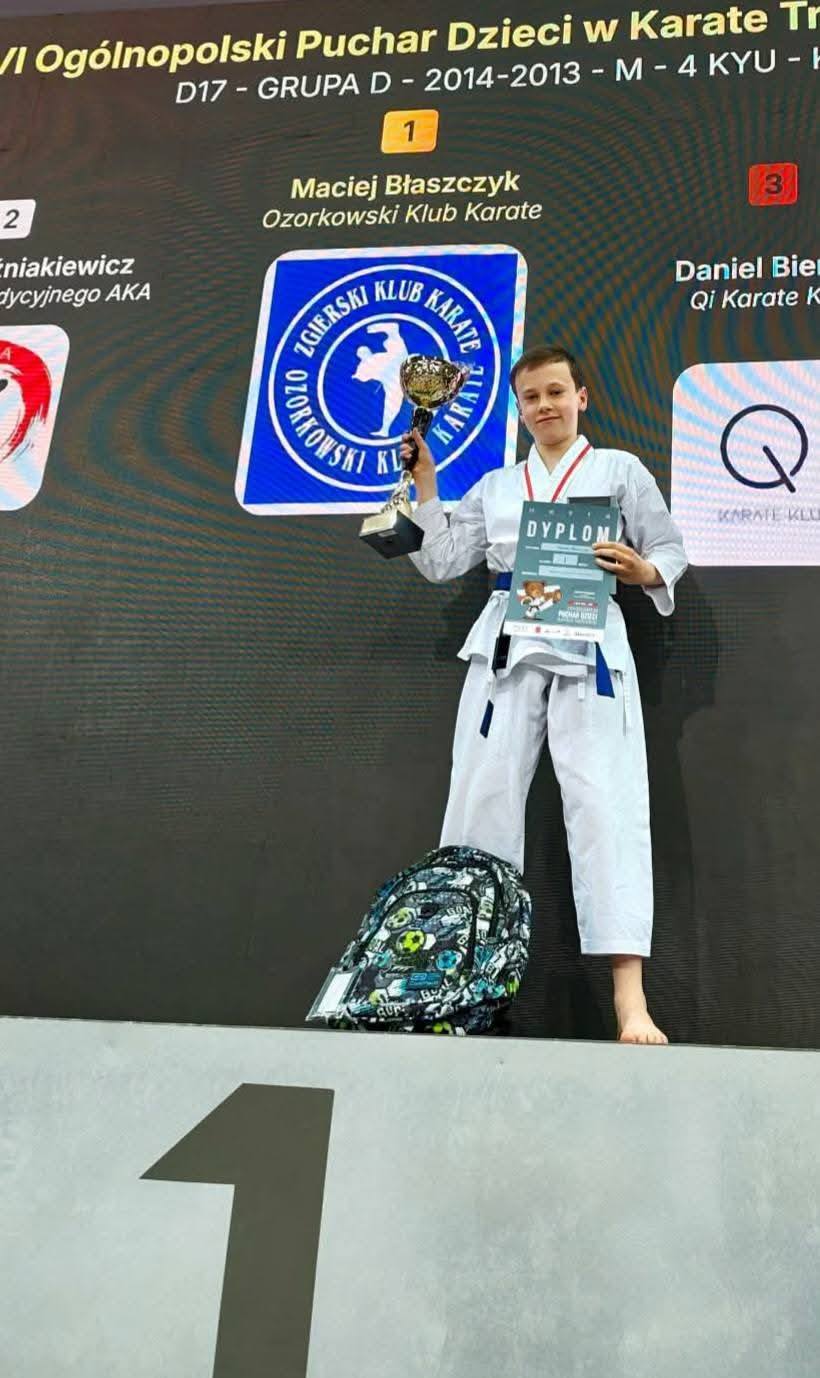 Uczeń w&nbsp;stroju karateki stoi na&nbsp;tle baner&oacute;w z&nbsp;informujących o&nbsp;turnieju i&nbsp;sponsorach pucharu Polski w&nbsp;karate. W prawej ręce trzyma puchar, a&nbsp;w lewej dyplom