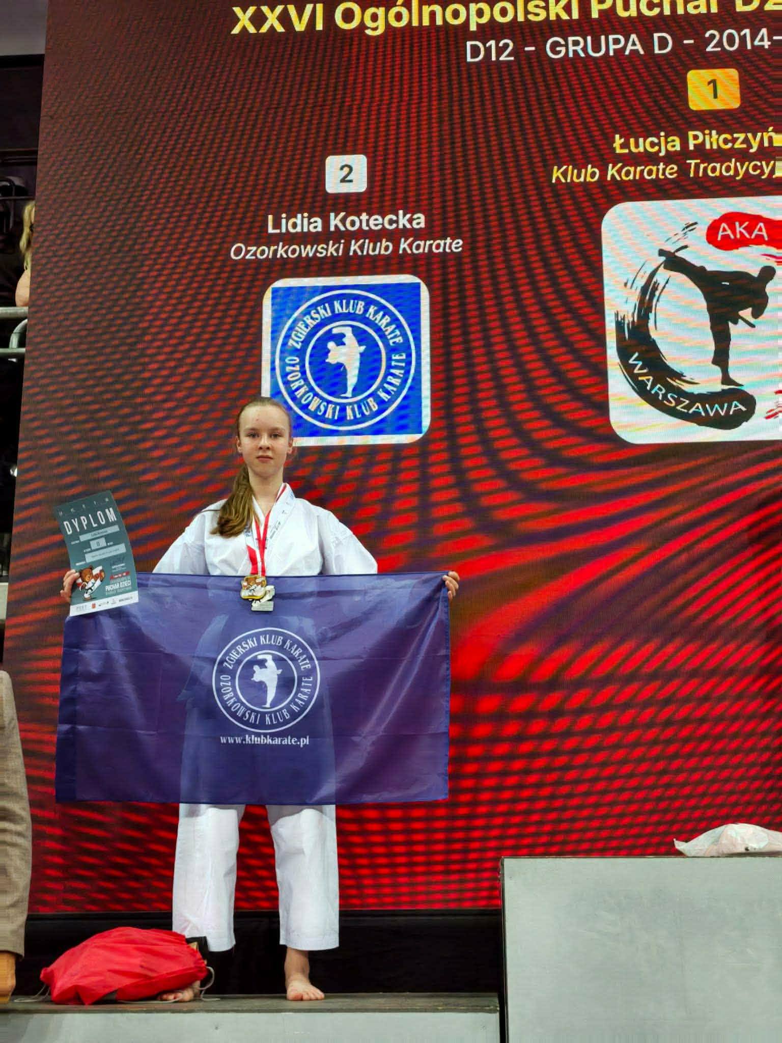 Uczeń w&nbsp;stroju karateki stoi na&nbsp;tle baner&oacute;w z&nbsp;informujących o&nbsp;turnieju i&nbsp;sponsorach pucharu Polski w&nbsp;karate. Przed sobą trzyma flagę klubu sportowego z&nbsp;Ozorkowa